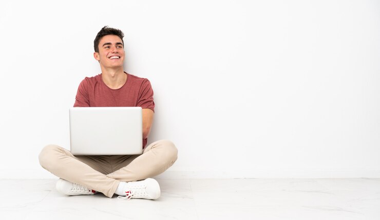 Foto hombre adolescente sentado en la flor con su computadora portátil mirando hacia arriba mientras sonríe