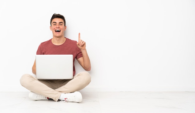 Hombre adolescente sentado en la flor con su computadora portátil pensando en una idea apuntando el dedo hacia arriba