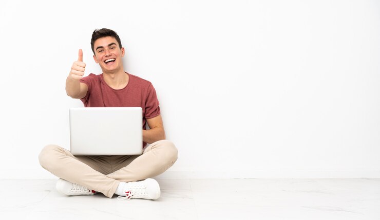Foto hombre adolescente sentado en la flor con su computadora portátil con los pulgares hacia arriba porque algo bueno ha sucedido