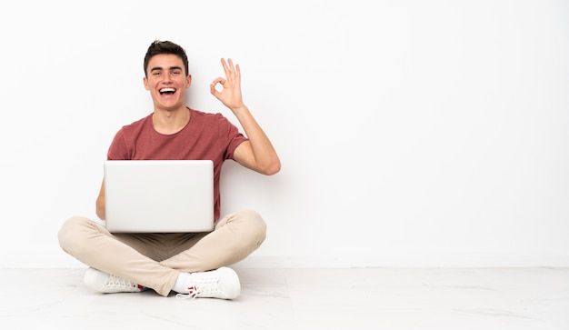 Foto hombre adolescente sentado en la flor con su computadora portátil sorprendido y mostrando signo ok
