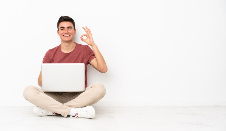 Foto hombre adolescente sentado en el suelo con su computadora portátil que muestra bien firmar con los dedos
