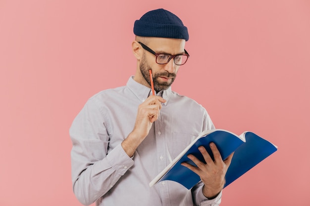 Hombre adulto sin afeitar concentrado sostiene un libro de texto azul y un lápiz, lee la información necesaria