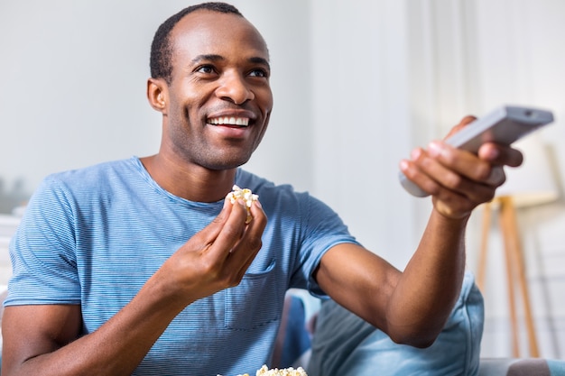 Hombre adulto alegre feliz sosteniendo un control remoto y comiendo palomitas de maíz mientras elige un canal de televisión