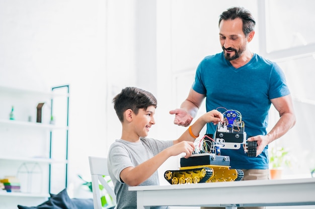 Hombre adulto alegre que sostiene un dispositivo robótico mientras ayuda a su hijo con el proyecto de ingeniería