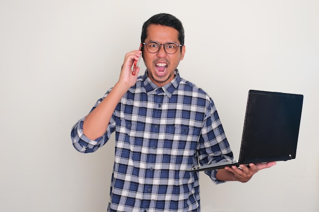 Foto hombre adulto asiático gritando enojado mientras contesta una llamada telefónica y sostiene una computadora portátil