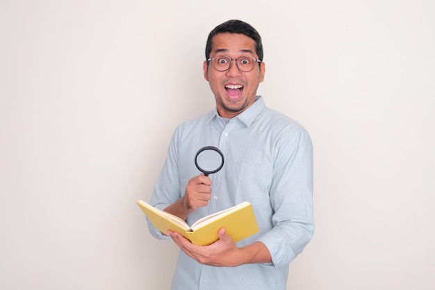 Hombre adulto asiático mostrando una expresión facial de wow mientras sostiene un libro y una lupa