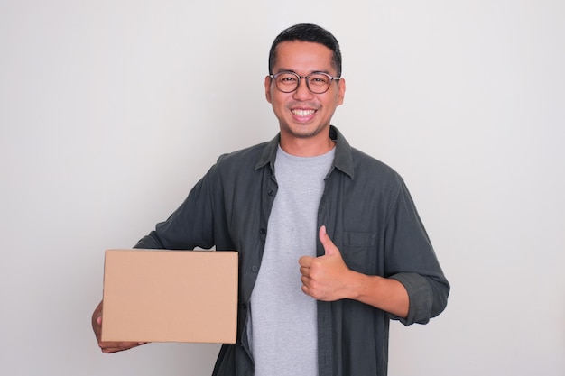 Foto hombre adulto asiático sonriendo y dando el pulgar hacia arriba mientras sostiene una caja de paquete