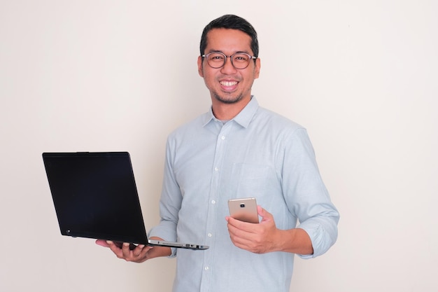 Hombre adulto asiático sonriendo feliz mientras sostiene una computadora portátil y un teléfono móvil