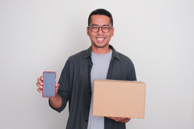 Foto hombre adulto asiático sonriendo mientras sostiene una caja de paquetes y muestra una pantalla de teléfono en blanco