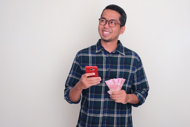 Foto hombre adulto asiático sonriendo y mirando al lado derecho mientras sostiene el teléfono y el dinero