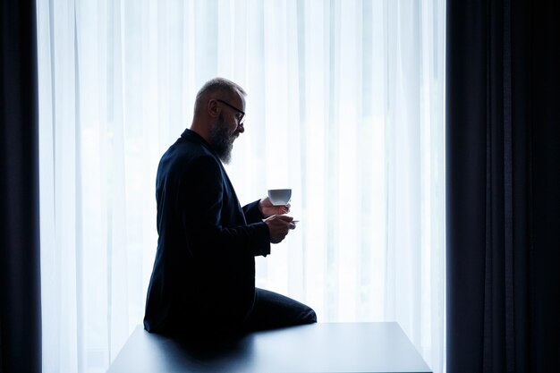 Un hombre adulto con barba bebe café y mira por la ventana. Concepto de recreación de campo de día de trabajo