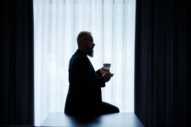 Un hombre adulto con barba bebe café y mira por la ventana. Concepto de recreación de campo de día de trabajo