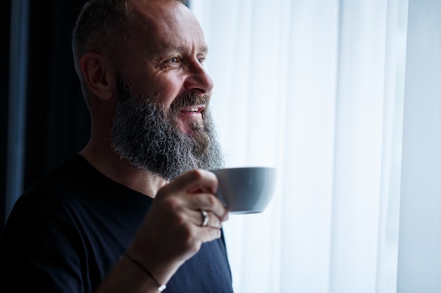 Un hombre adulto con barba bebe café y mira por la ventana. Concepto de recreación de campo de día de trabajo