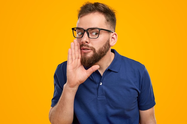 Hombre adulto con barba en camisa azul y gafas manteniendo la mano cerca de la boca mientras revela información confidencial sobre fondo amarillo