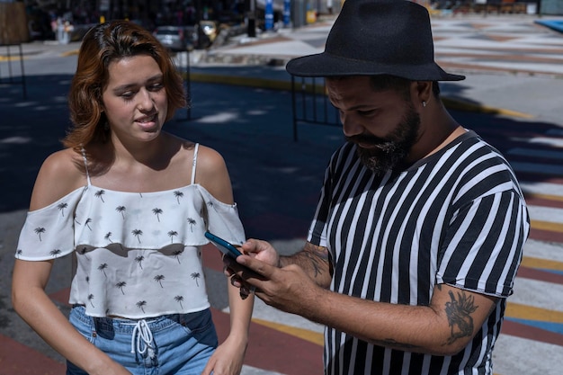 Hombre adulto con barba y sombrero con una joven latinoamericana caminando por una ciudad de vacaciones usando el mapa de su celular para guiarlo Concepto de vacaciones