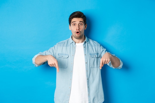 Hombre adulto barbudo sorprendido en traje casual, señalando con el dedo hacia abajo y mirando sorprendido, haciendo preguntas sobre promoción, de pie sobre una pared azul