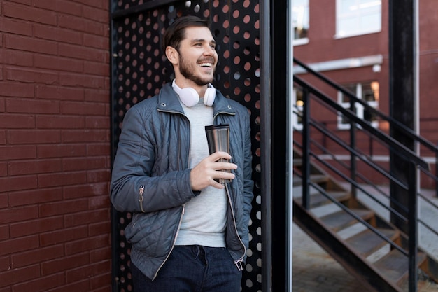 Hombre adulto bebiendo café de una taza reutilizable en la calle con auriculares alrededor del cuello junto a