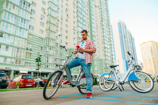 Hombre adulto con camisa y barba se sienta en una bicicleta shering y usa un teléfono inteligente El turista alquila una bicicleta a través de una aplicación móvil Transporte ecológico