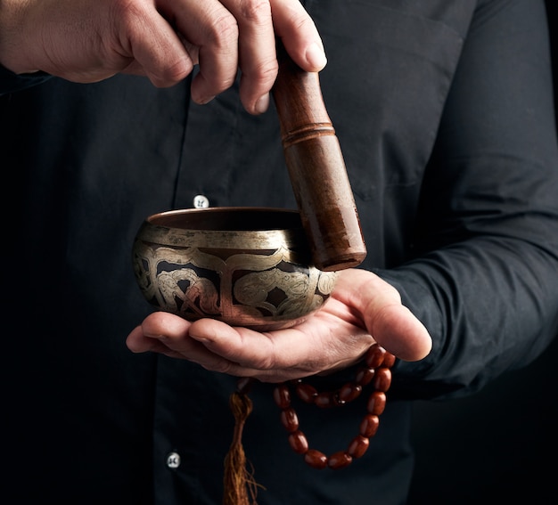 Un hombre adulto con una camisa negra gira un palo de madera alrededor de un cuenco de agua tibetano de cobre. ritual de meditación