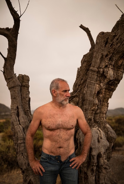 Foto hombre adulto sin camisa de pie junto al tronco desnudo del árbol de olivo contra el cielo despejado