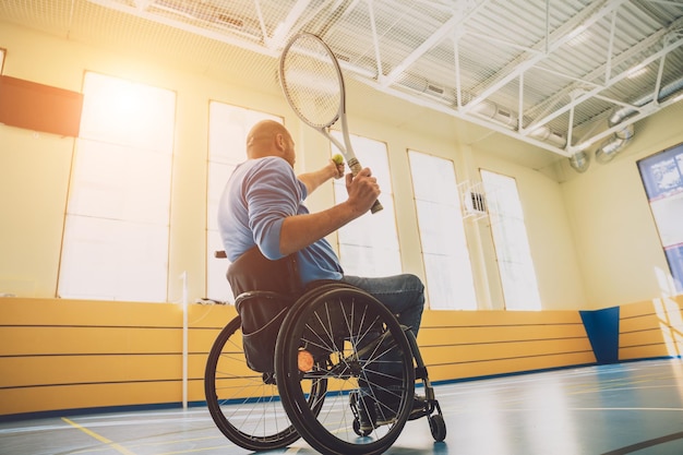 Hombre adulto con discapacidad física que usa silla de ruedas jugando al tenis en una cancha de tenis cubierta