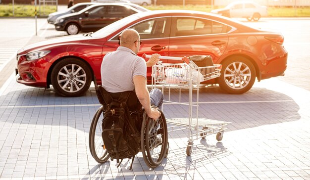 Hombre adulto discapacitado en silla de ruedas empuja un carro hacia un automóvil en el estacionamiento de un supermercado