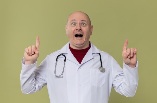 Hombre adulto emocionado en uniforme médico con estetoscopio apuntando hacia arriba