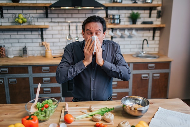 Hombre adulto enfermo estornudando a servilleta blanca. Se para a la mesa en la cocina. Escritorio lleno de coloridas verduras y especias saludables.