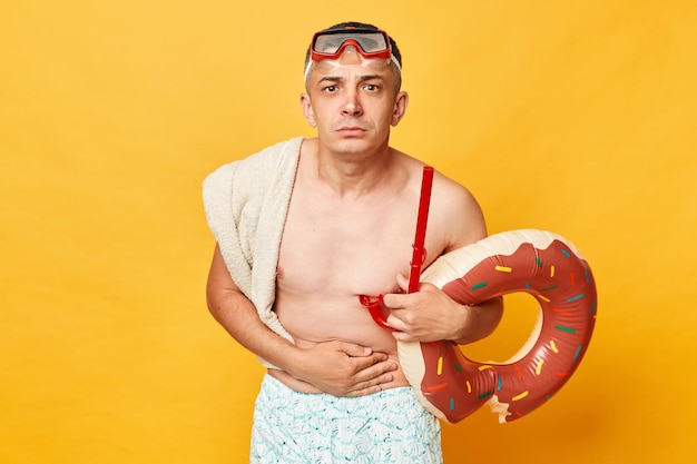 Hombre adulto enfermo y malsano que usa pantalones cortos, traje de baño, gafas de esnórquel, sostiene un anillo de goma de donut y una bolsa aislada en el fondo amarillo, siente dolor en la cara fruncida del vientre