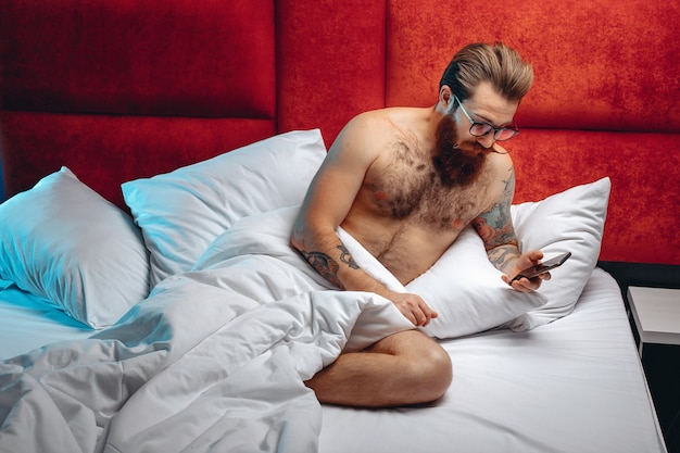 Hombre adulto enfocado sexual con gafas y sentado en una cama blanca mirando el teléfono antes de irse a la cama.