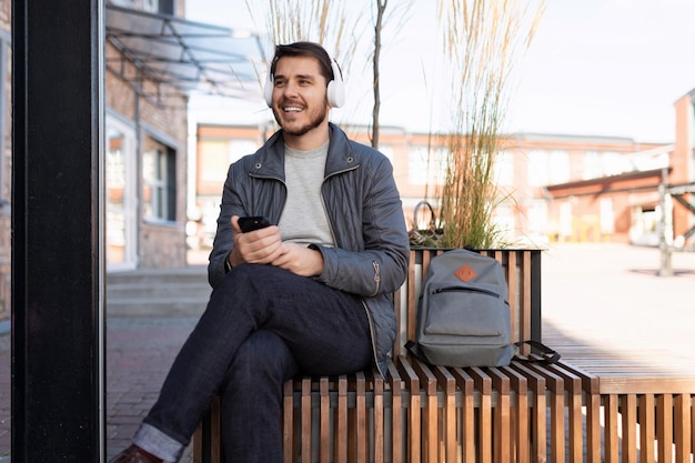 Hombre adulto escuchando música con auriculares con un teléfono en las manos sentado en un banco junto a un