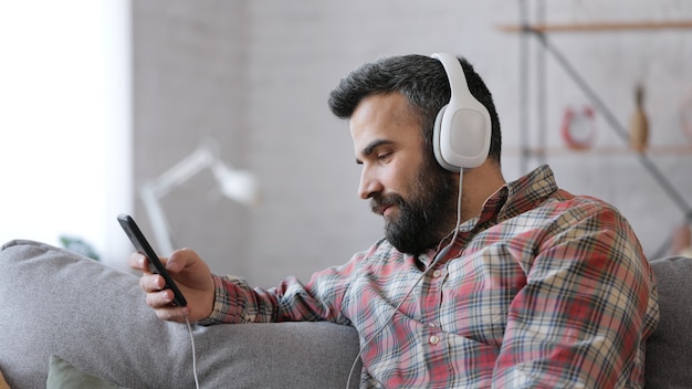 Hombre adulto feliz en auriculares blancos disfruta de escuchar música favorita en el teléfono inteligente con la aplicación de música.