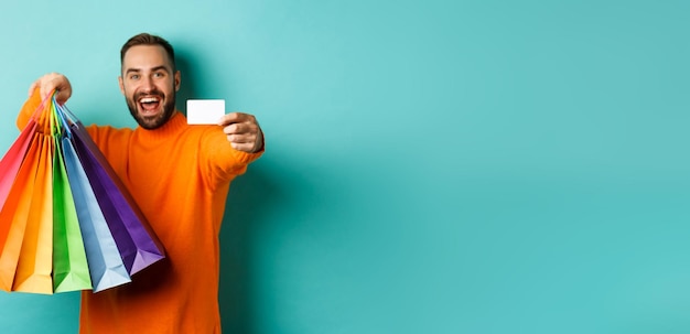 Foto hombre adulto feliz mostrando tarjeta de crédito y bolsas de compras de pie contra el fondo turquesa