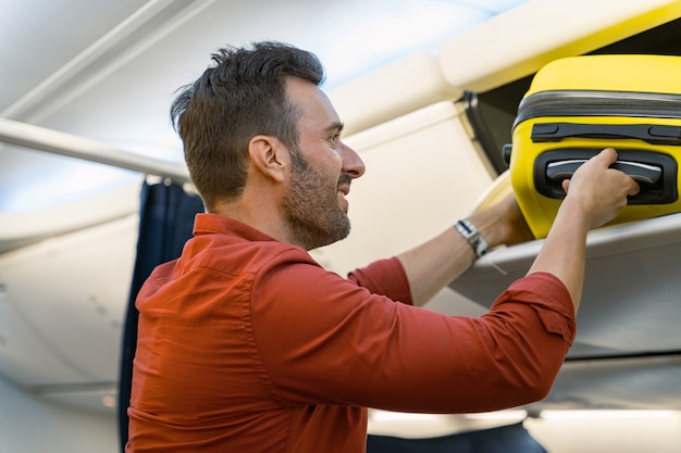 Hombre adulto feliz poniendo una maleta en un estante en un avión