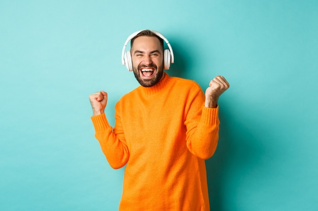 Hombre adulto feliz en suéter naranja, mirando hacia arriba y escuchando música en auriculares, de pie sobre fondo azul.