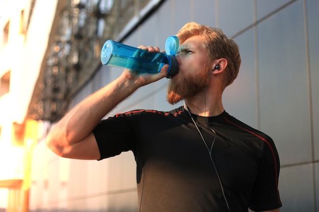 Hombre adulto guapo bebiendo agua de la botella de fitness mientras está parado afuera, al atardecer o al amanecer. Corredor