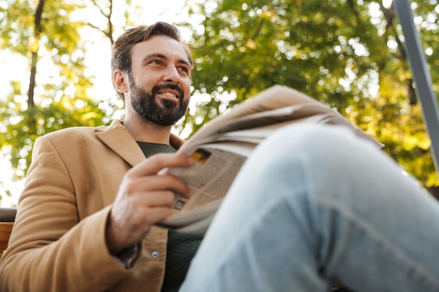 Hombre adulto guapo en chaqueta sonriendo y leyendo el periódico mientras está sentado en un banco en el parque