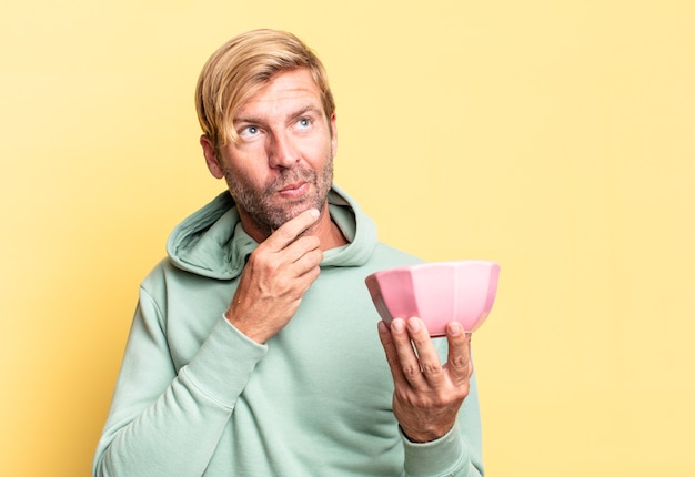 Hombre adulto guapo rubio sosteniendo una olla vacía contra la pared amarilla