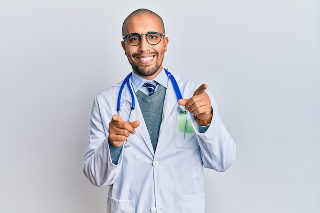 Hombre adulto hispano vestido con uniforme médico y estetoscopio señalando con el dedo a la cámara con cara feliz y divertida buena energía y vibraciones