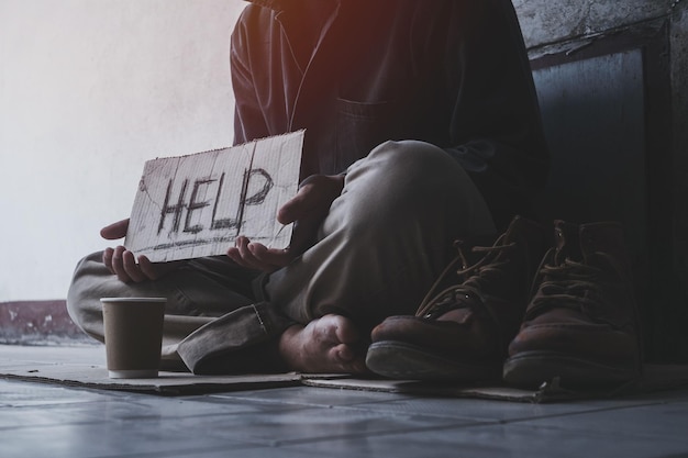 Foto hombre adulto sin hogar sentado en la calle a la sombra del edificio y pidiendo ayuda y dinero con el cartel. concepto de personas sin hogar.