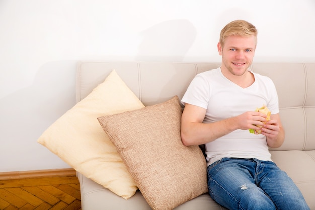 Un hombre adulto joven comiendo un sándwich en casa en el sofá.