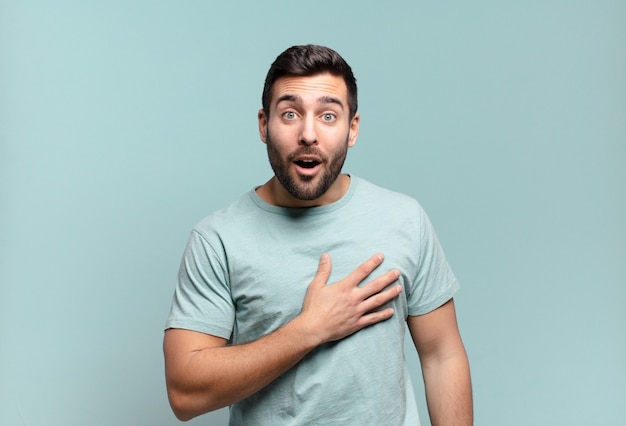 Hombre adulto joven guapo que se siente consternado y sorprendido, sonriendo, tomando la mano al corazón, feliz de ser el indicado o mostrando gratitud