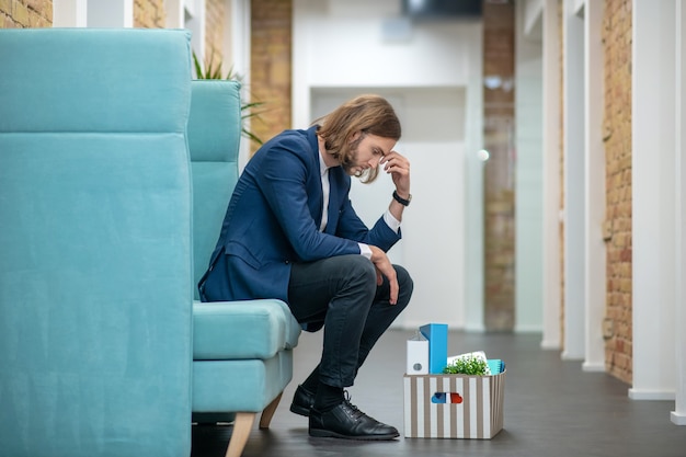 Hombre adulto joven infeliz en traje de negocios sentado en el sofá en el pasillo de la oficina triste preocupante
