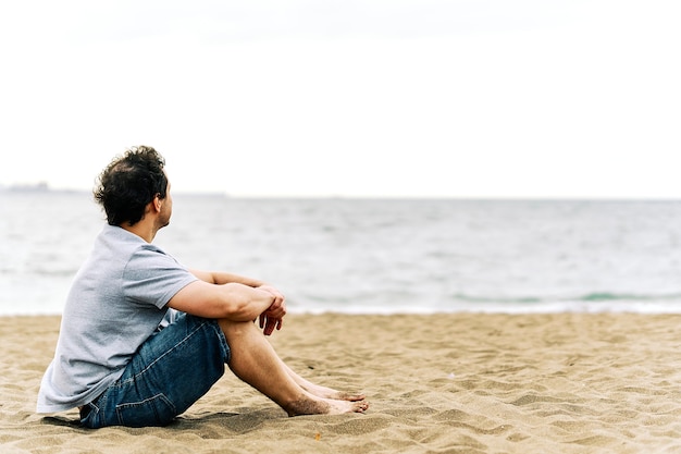 Hombre adulto joven pensativo sentado en la arena de la playa con gesto melancólico