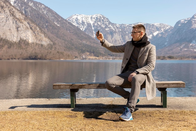 Hombre adulto joven sentado al aire libre en el banco y haciendo selfie con su teléfono inteligente disfrutando de las montañas lago buen tiempo cielo azul sol Hermoso paisaje increíble Tiempo de viaje de vacaciones