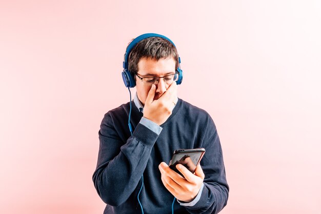 Hombre adulto joven de treinta y cinco años en camisa y suéter azul con cuello en v con fondo rosa con gafas, auriculares y smartphone, tapándose el rostro preocupado