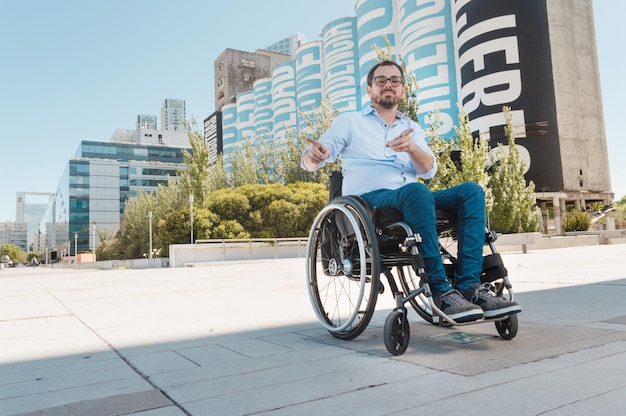 Hombre adulto latino hispano caucásico, con barba y anteojos, sonriendo feliz en una silla de ruedas animando a señalar la cámara con el dedo, al aire libre.