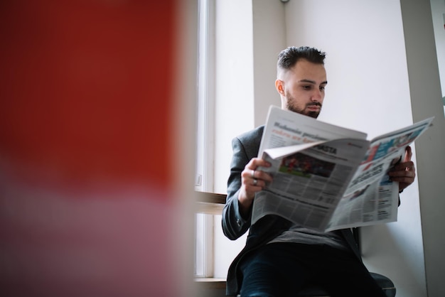 Hombre adulto leyendo el periódico mientras está sentado en el alféizar de la ventana