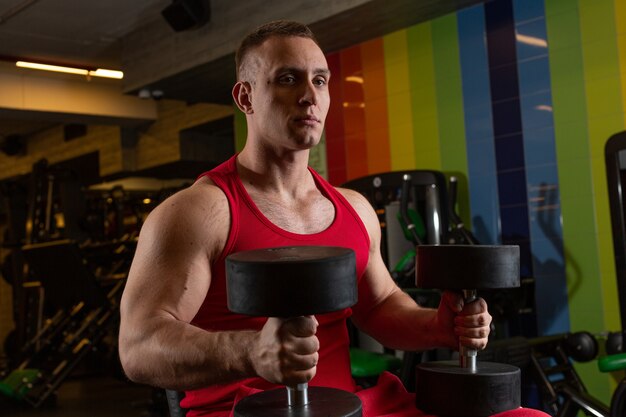 Un hombre adulto luciendo un físico se sienta en un simulador de gimnasio sosteniendo pesas y mirando con nostalgia hacia adelante.