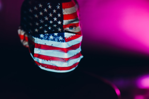 Hombre adulto en mascarilla médica con la bandera de Estados Unidos en su rostro en la oscuridad.